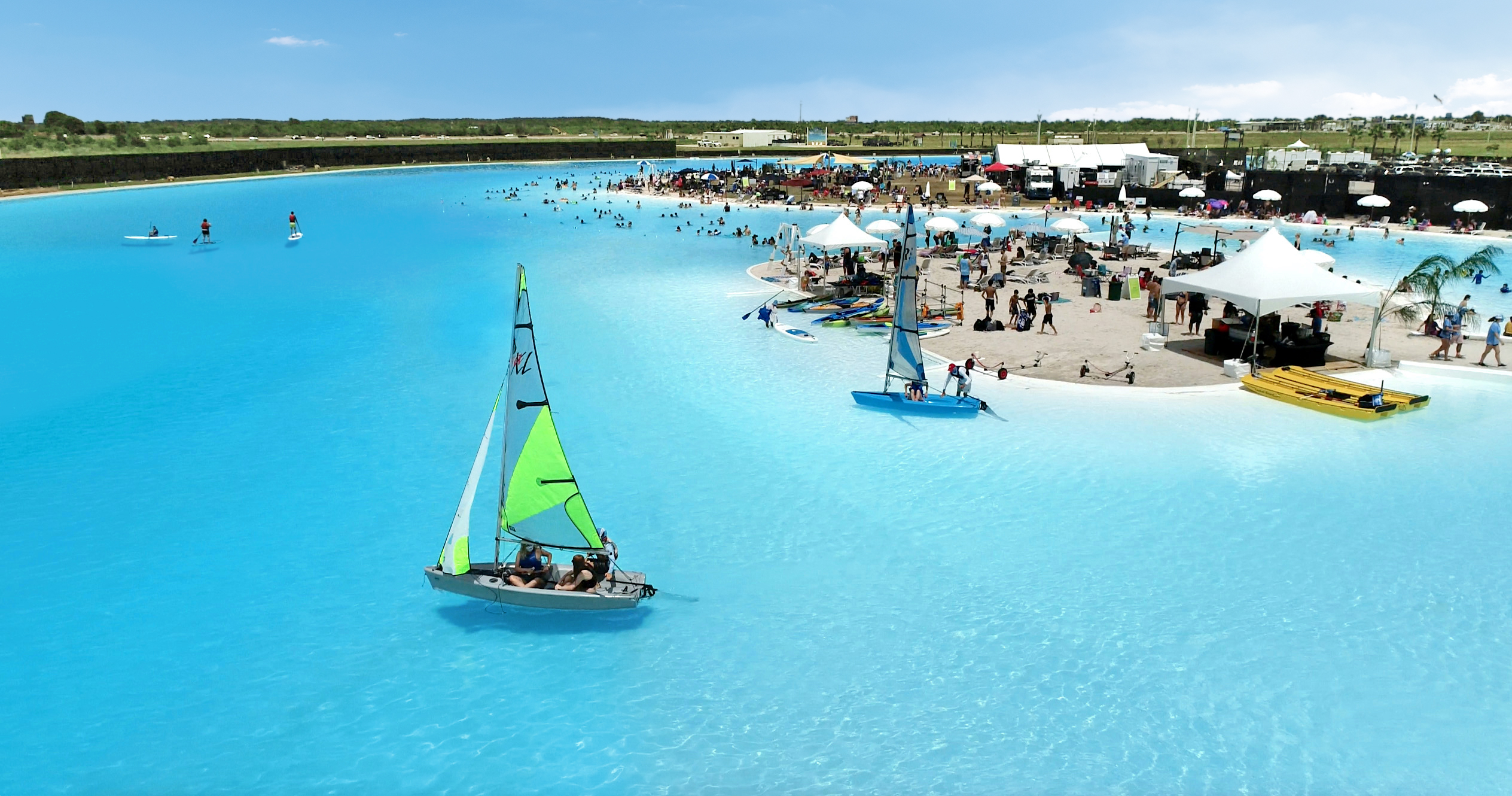 Aerial view of a large-scale turqoiuse lagoon in US, with a small sailing boat with a green sail and hundreds of people swimming