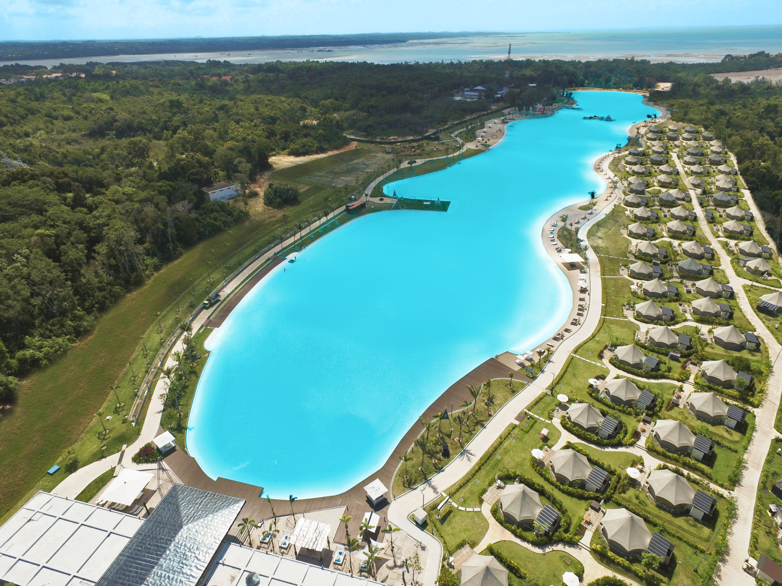 Aerial view of Southeast Asia's largest crystalline lagoon in Bintan, powered by Crystal Lagoons technology. Dozens of glamping huts surround the beach